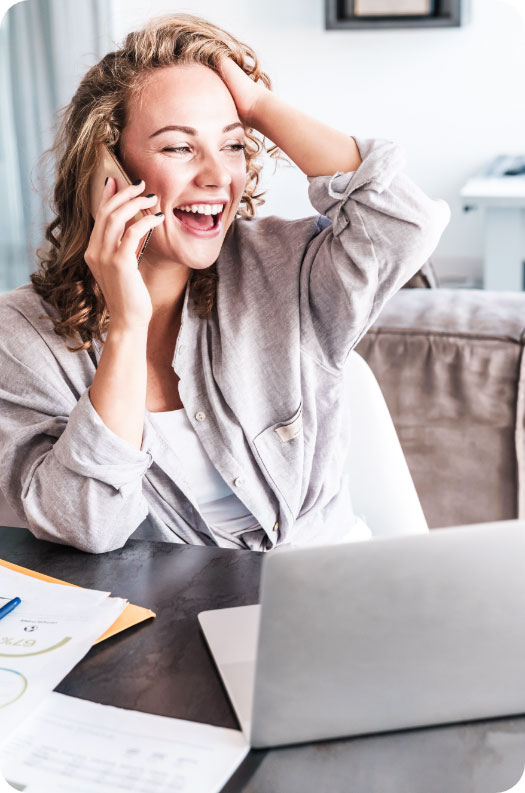 Eine strahlende Frau, die vor einem Laptop sitzt, freut sich gerade über die telefonische Nachricht über ihren neuen Job, den sie dank Weiterbildung bekommen hat.